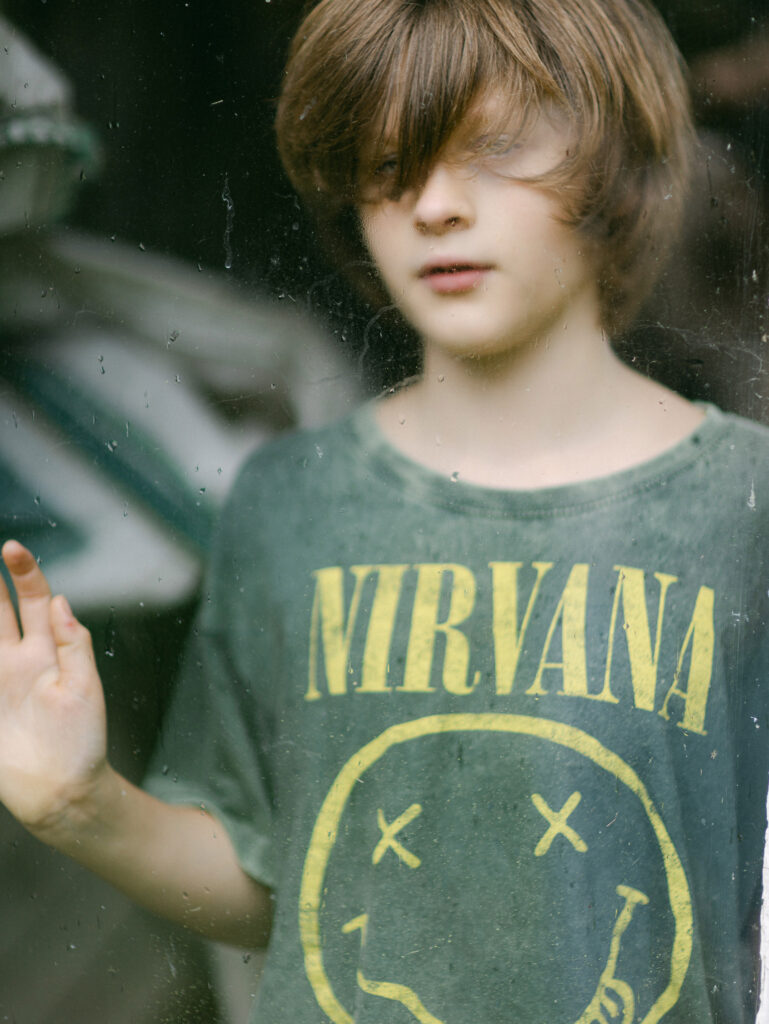Boy with long hair with his head squashed against a window looking bored but playful wearing a Nirvana t shirt